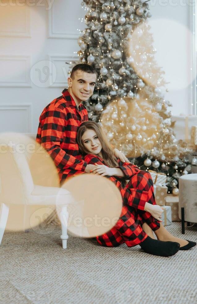 Jeune magnifique couple dans correspondant à rouge pyjamas près le Noël arbre. joie, câlins et bisous. Nouveau années vacances et cadeaux en dessous de le Noël arbre photo