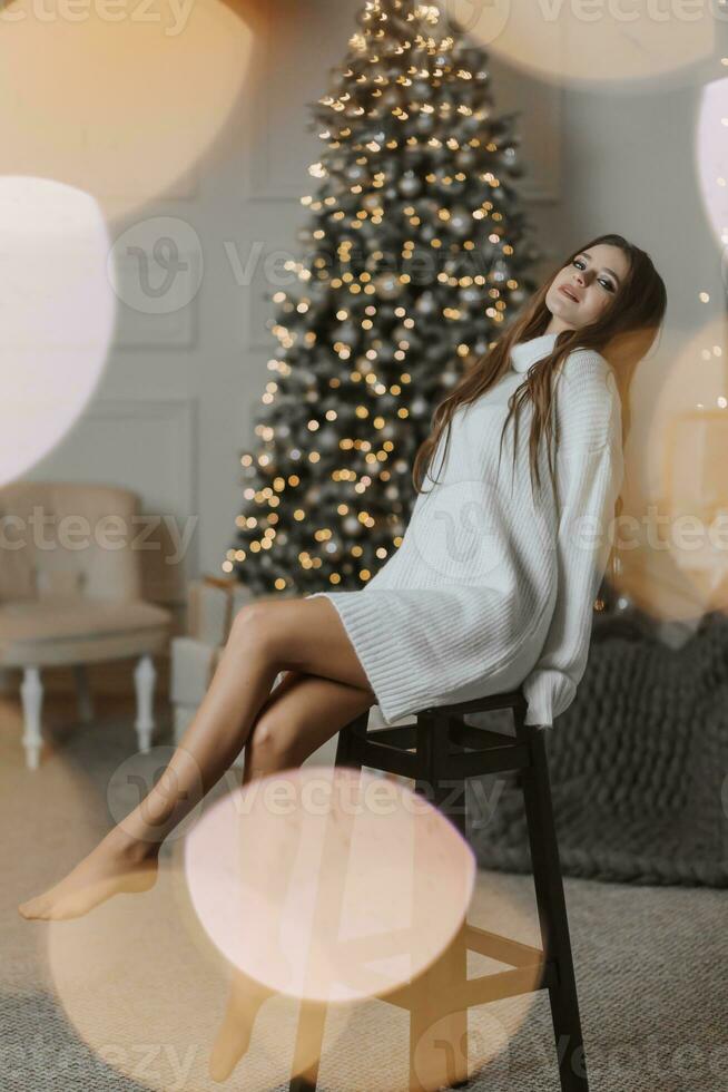 une magnifique et élégant Jeune fille dans une blanc chandail pose dans le Noël lumières sur le Contexte de le Noël arbre photo