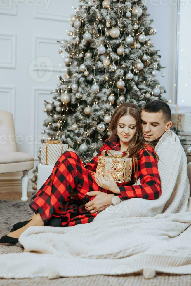 Jeune magnifique famille couple dans identique rouge pyjamas près le Noël arbre, enveloppé dans une couverture, réjouir et échange Nouveau années cadeaux. Nouveau années vacances et cadeaux en dessous de le Noël arbre photo