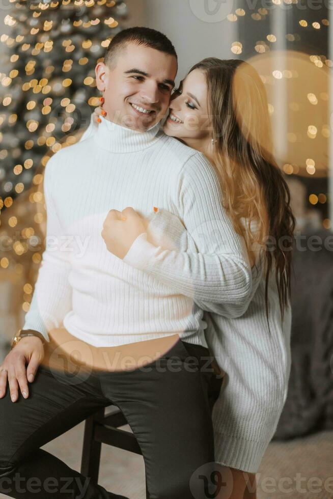 une magnifique et élégant Jeune famille dans blanc chandails étreinte et réjouir dans le Noël lumières contre le Contexte de le Noël arbre. Nouveau année concept photo