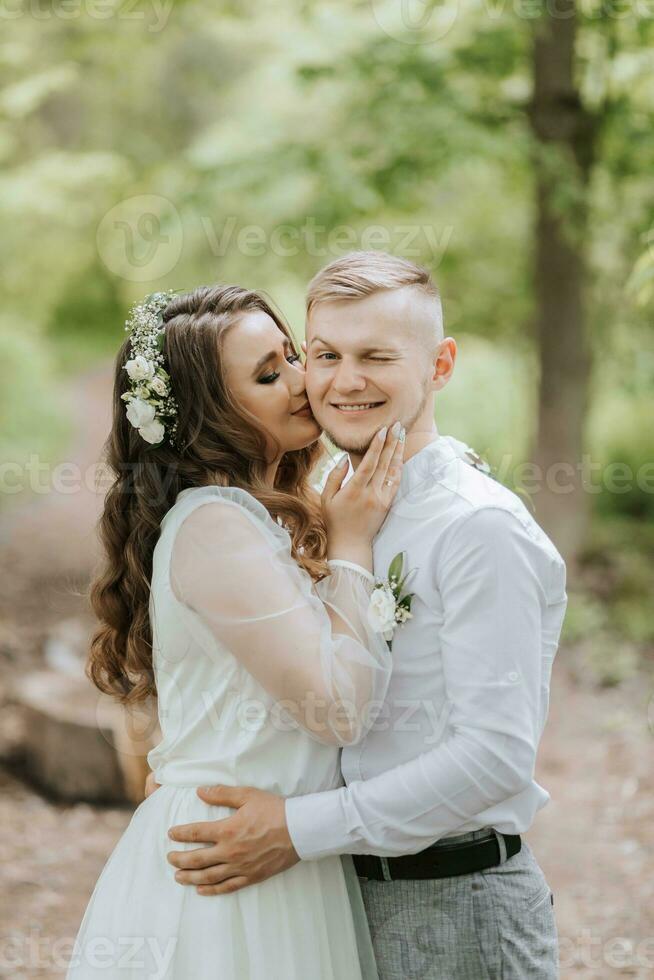 mariage marcher dans le forêt. portrait de une couple dans l'amour dans le forêt, le la mariée baisers le jeune marié sur le joue, il sourit photo