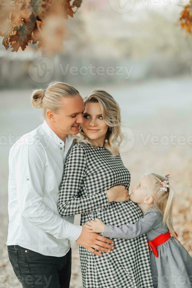 portrait de une Jeune Enceinte femme avec sa famille à l'extérieur dans une parc parmi jauni feuilles photo