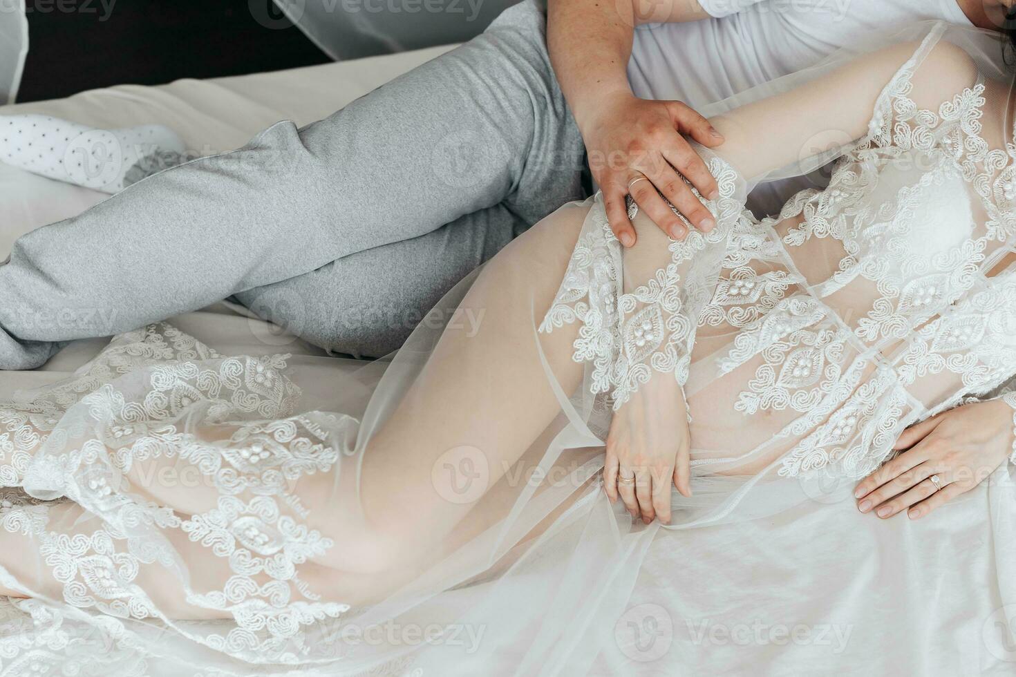 proche en haut photo de Jeune brunette Enceinte femme dans sa pièce dans blanc dentelle robe sur lit avec homme. maternité. concept de grossesse, maternité, préparation et en attendant.