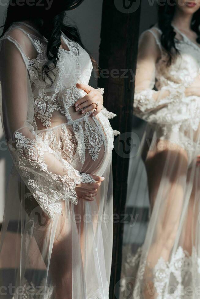 une magnifique Enceinte femme dans une dentelle transparent robe câlins sa ventre avec sa mains près le miroir. concept de grossesse, maternité, préparation et en attendant. le beauté de une femme pendant grossesse photo