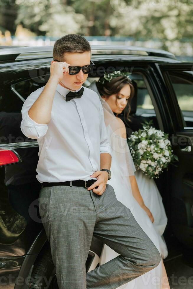 mariage portrait, photo de une élégant jeune marié dans une blanc chemise et arc attacher et une brunette la mariée avec une bouquet de fleurs près une noir auto.