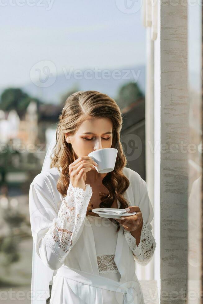 une magnifique la mariée dans une blanc peignoir les boissons café de une blanc tasse sur une Hôtel balcon surplombant une gros ville. sur votre mariage journée avant mariage. photo