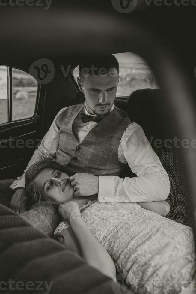 une luxueux et élégant mariage couple tendrement embrasse dans le milieu de une élégant rétro auto. le photo a été pris par le pare-brise. magnifique la mariée et Beau jeune marié dans rétro style.