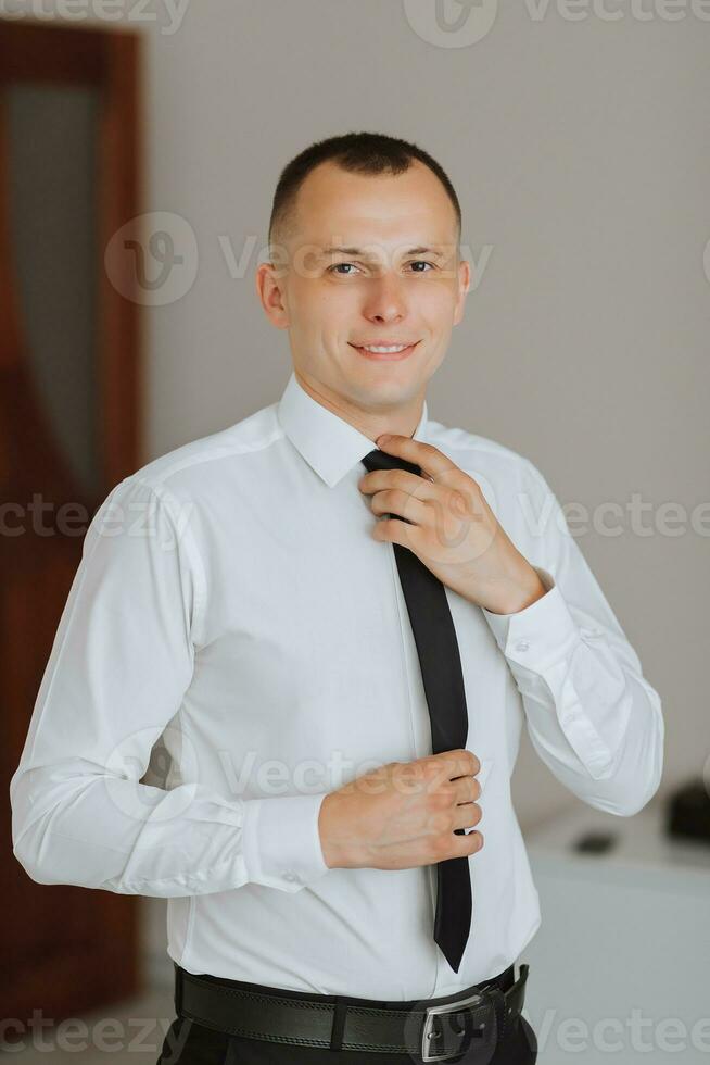 Beau souriant et élégant jeune marié dans une blanc chemise avec une attacher dans le sien mains, en train de préparer pour le mariage photo