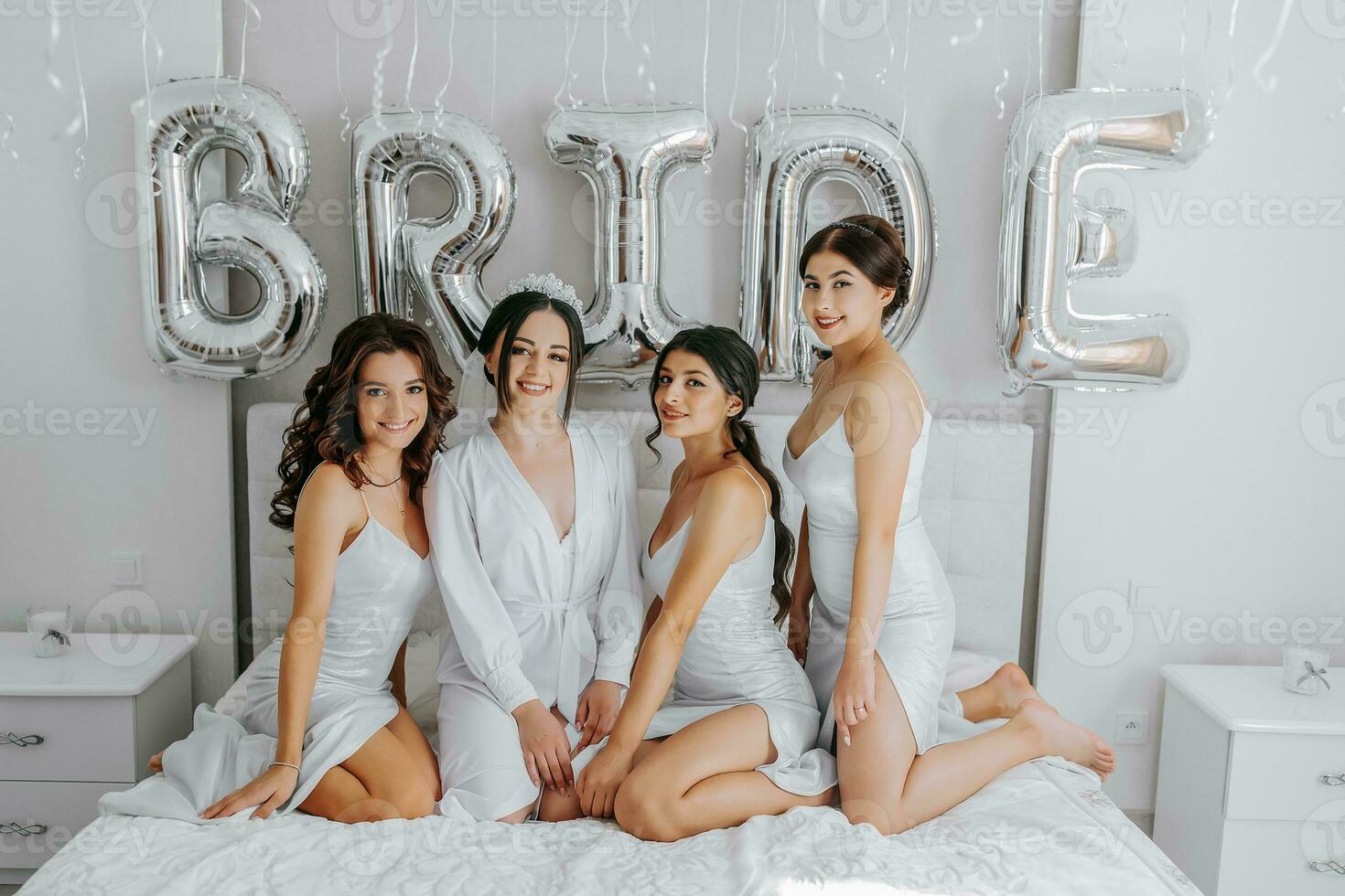 Jeune demoiselles d'honneur dans blanc soie Robes sur une lit dans le de la mariée chambre. magnifique femmes célébrer bachelorette fête séance sur lit. photo