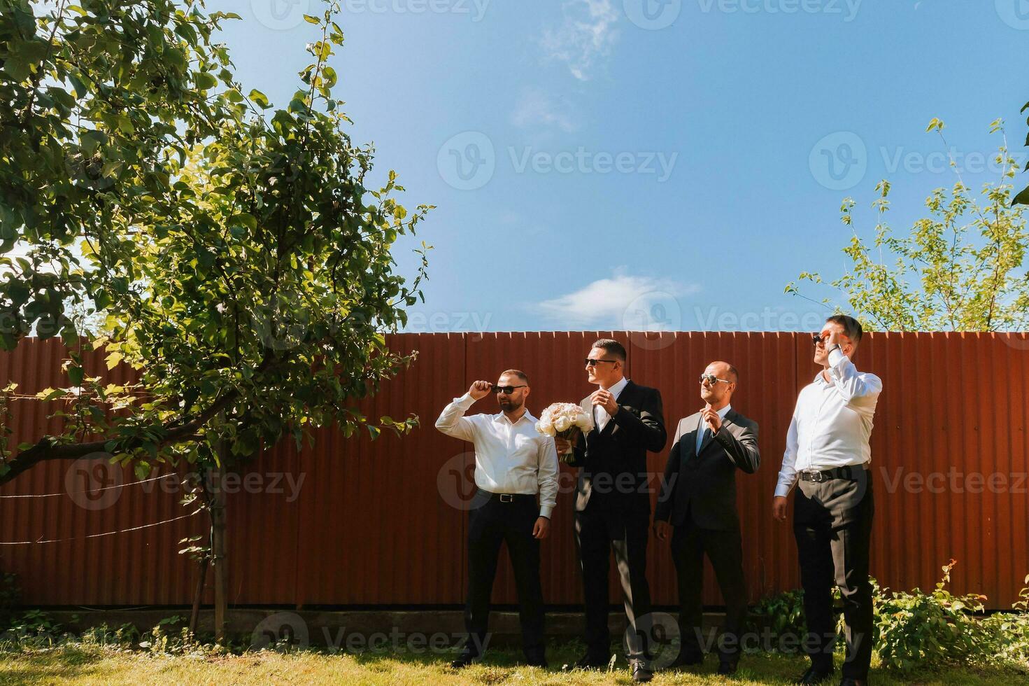 le jeune marié dans une noir costume et des lunettes et le sien élégant copains portant blanc chemises et noir un pantalon et des lunettes sont permanent dans le arrière-cour dans le jardin. le jeune marié est en portant une bouquet. photo