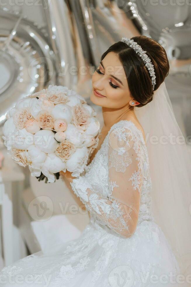 une magnifique Jeune la mariée dans une mariage robe avec une bouquet de pivoines dans sa mains est séance sur une lit dans une chambre. final les préparatifs pour le mariage. le la mariée est attendre pour le jeune marié. matin, la mariée. photo