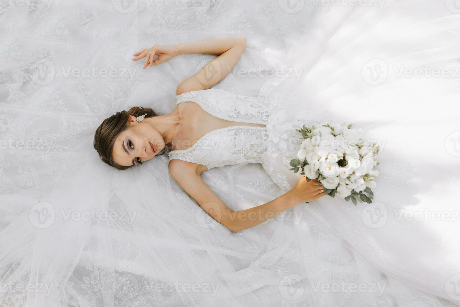 magnifique la mariée avec mariage bouquet de fleurs, attrayant femme dans mariage robe. le la mariée rêveusement mensonges sur sa robe, le photo est pris de au dessus