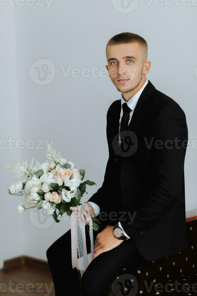 portrait. une homme dans une blanc chemise, une noir costume est séance, posant sur une canapé, en portant une bouquet. une élégant montre. Pour des hommes style. mode. affaires photo