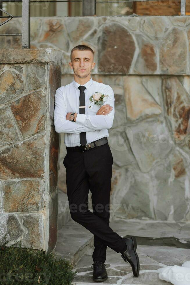 une homme dans noir un pantalon et une blanc chemise pose près une pierre mur. une élégant montre. Pour des hommes style. mode. entreprise. photo