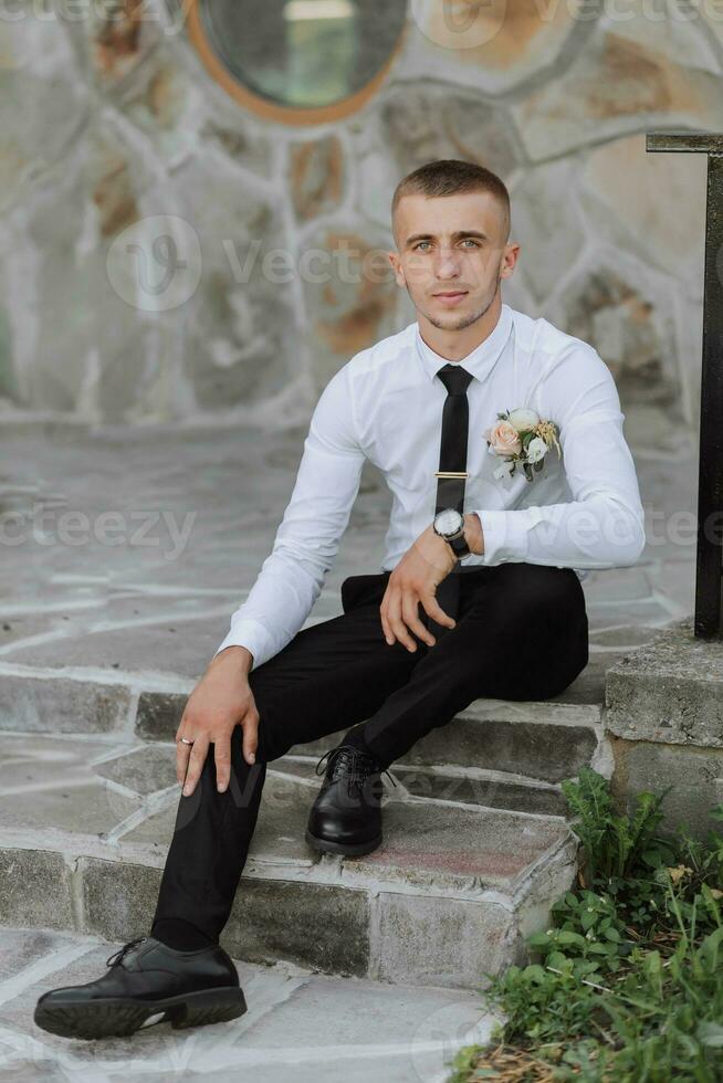 une homme dans noir un pantalon et une blanc chemise est assis et pose près une pierre mur. une élégant montre. Pour des hommes style. mode. entreprise. photo