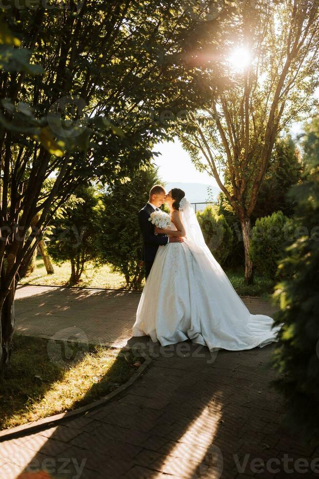 chaud ensoleillé portrait de une content la mariée avec une bouquet de orchidées dans sa mains et une jeune marié dans une classique costume, à le coucher du soleil. chaud été temps. une longue voile, une luxueux blanc robe avec une train. photo