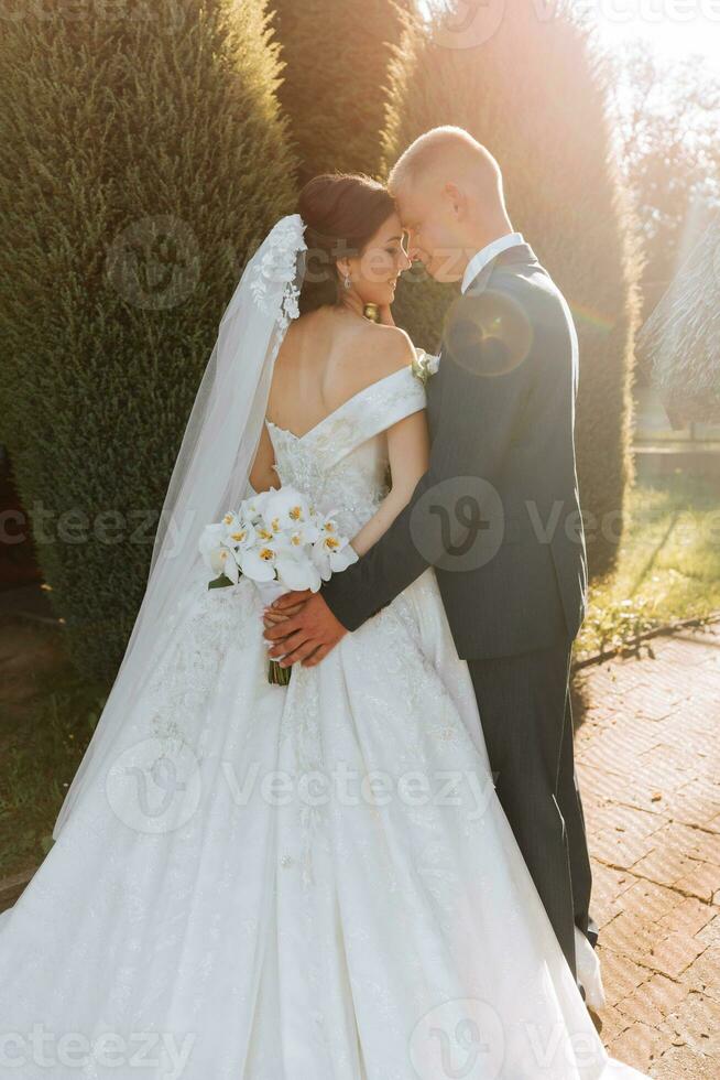 chaud ensoleillé portrait de une content la mariée avec une bouquet de orchidées dans sa mains et une jeune marié dans une classique costume, à le coucher du soleil. chaud été temps. une longue voile, une luxueux blanc robe avec une train. photo