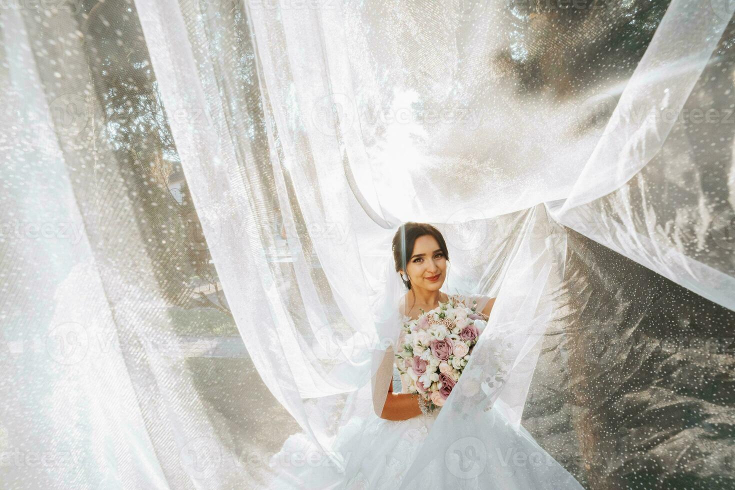 magnifique la mariée en dessous de une voile dans une à la mode mariage robe sur une Naturel Contexte dans le parc à le coucher du soleil. une étourdissant Jeune la mariée est incroyablement heureux. content fille sur sa mariage journée photo