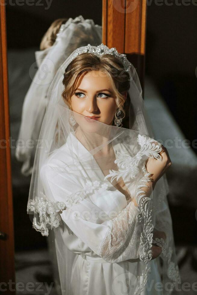 préparation pour le mariage. magnifique Jeune la mariée dans une blanc peignoir dans le chambre. une luxueux modèle près une miroir avec une voile et une tiare sur sa diriger. Matin de le la mariée photo