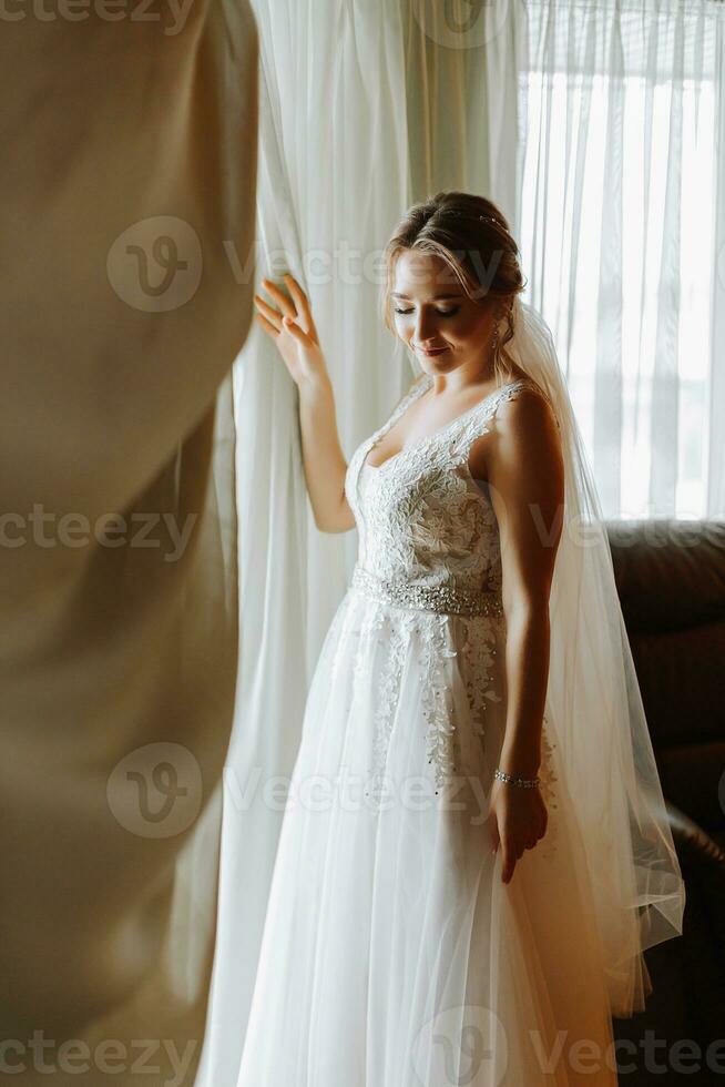 magnifique et doux la mariée dans une mariage robe dans le Matin posant près le fenêtre avec Naturel lumière. magnifique Naturel se maquiller. mariage coiffure. Matin de le la mariée. photo