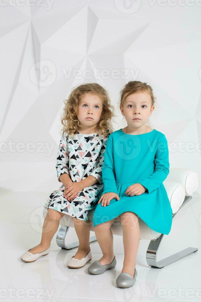 deux peu copines dans le identique Robes de différent couleurs séance sur une chaise dans une studio avec blanc des murs photo