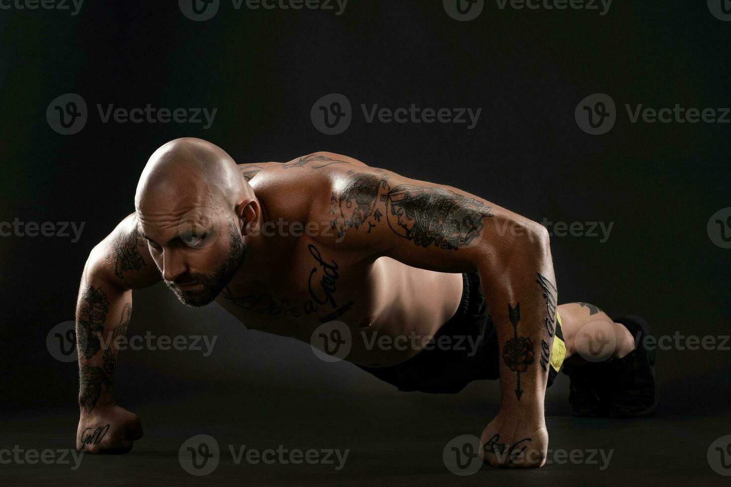 athlétique chauve, tatoué homme dans noir short et baskets est posant contre une noir Contexte. fermer portrait. photo