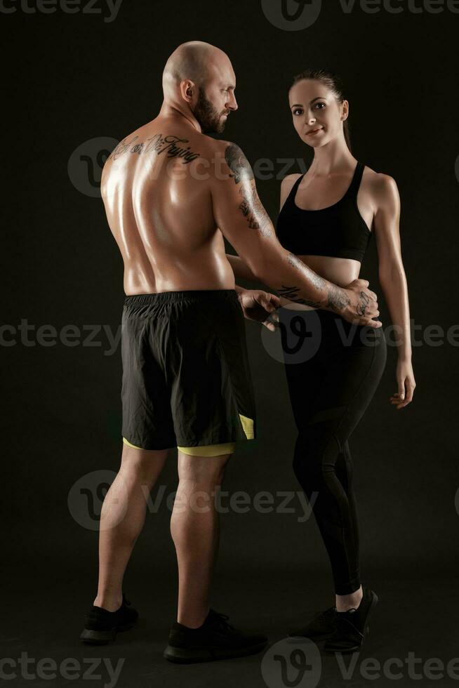 athlétique homme dans short et baskets avec brunette femme dans leggings et Haut posant sur noir Contexte. aptitude couple, Gym concept. photo