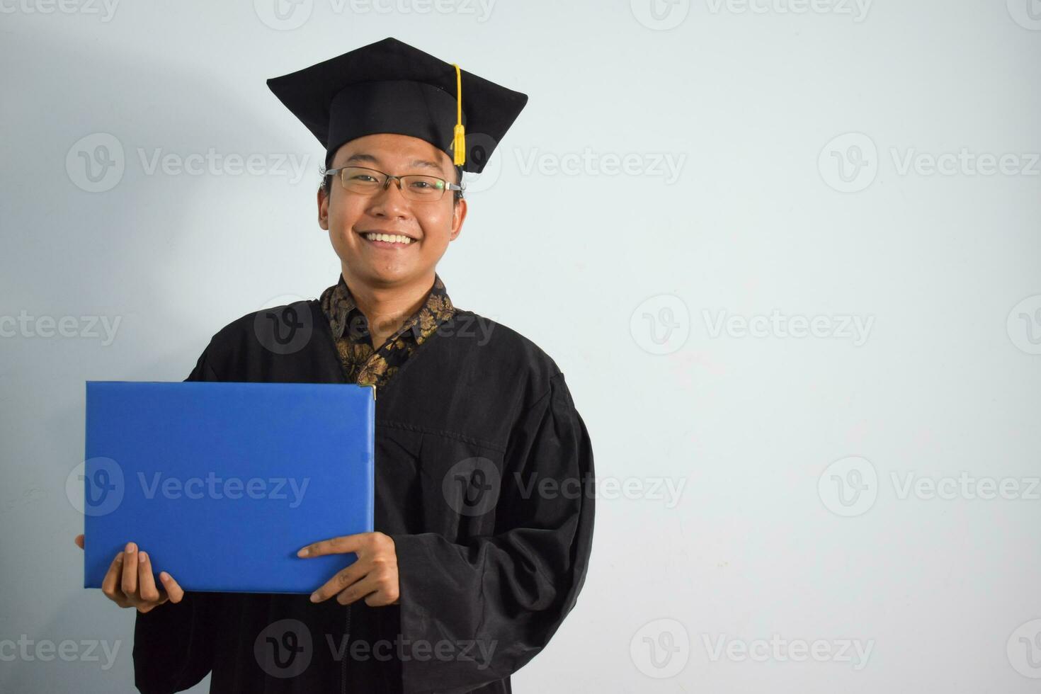 expressif de adulte Indonésie Masculin porter l'obtention du diplôme peignoir, chapeau et lunettes, asiatique Masculin l'obtention du diplôme apporter Vide bleu certificat isolé sur blanc arrière-plan, expressions de portrait l'obtention du diplôme photo