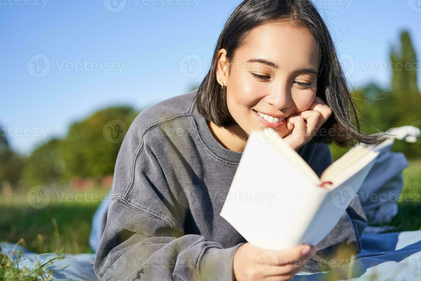 portrait de magnifique souriant asiatique fille, en train de lire dans parc, mensonge sur herbe avec préféré livre. loisir et gens concept photo