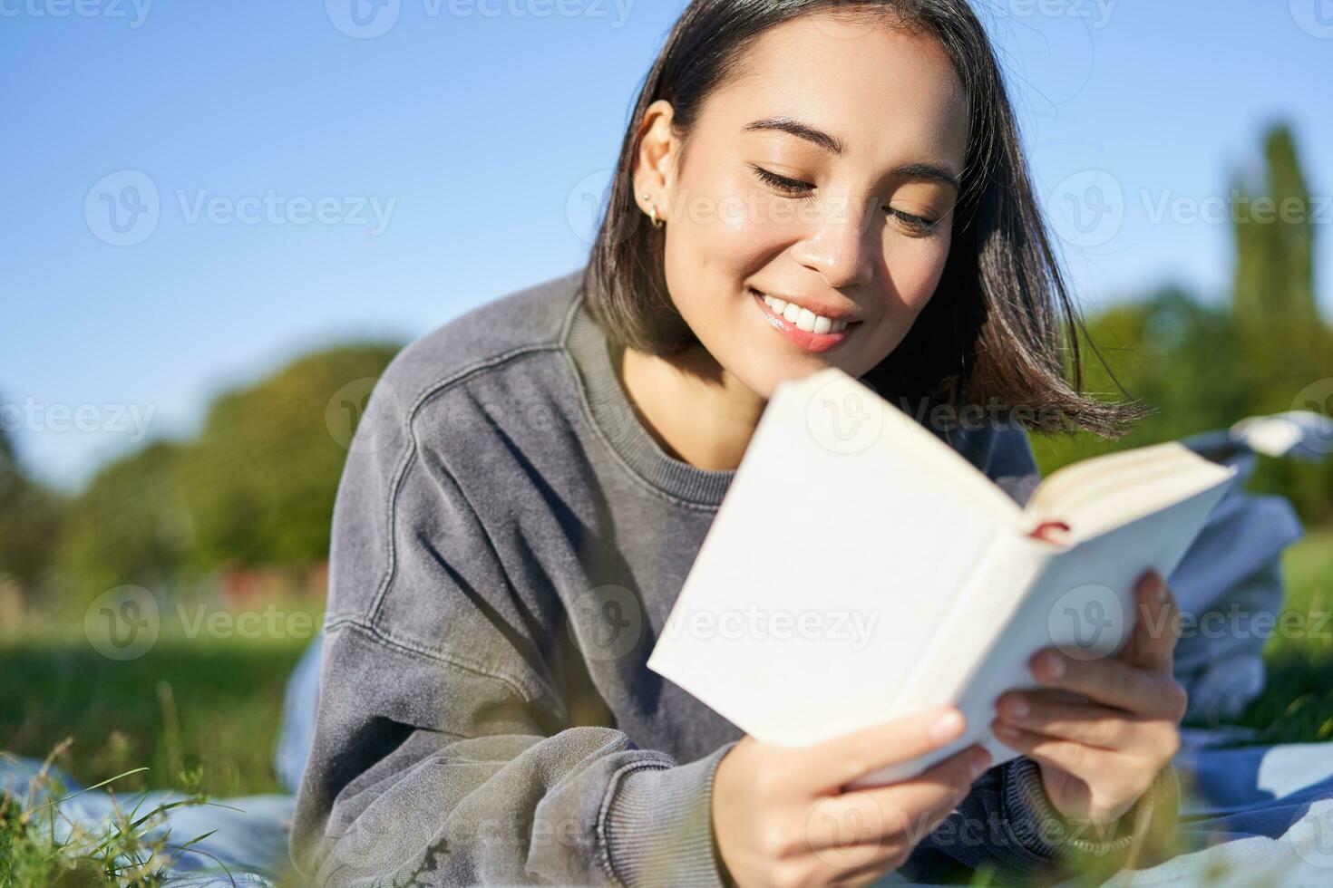 portrait de magnifique souriant asiatique fille, en train de lire dans parc, mensonge sur herbe avec préféré livre. loisir et gens concept photo