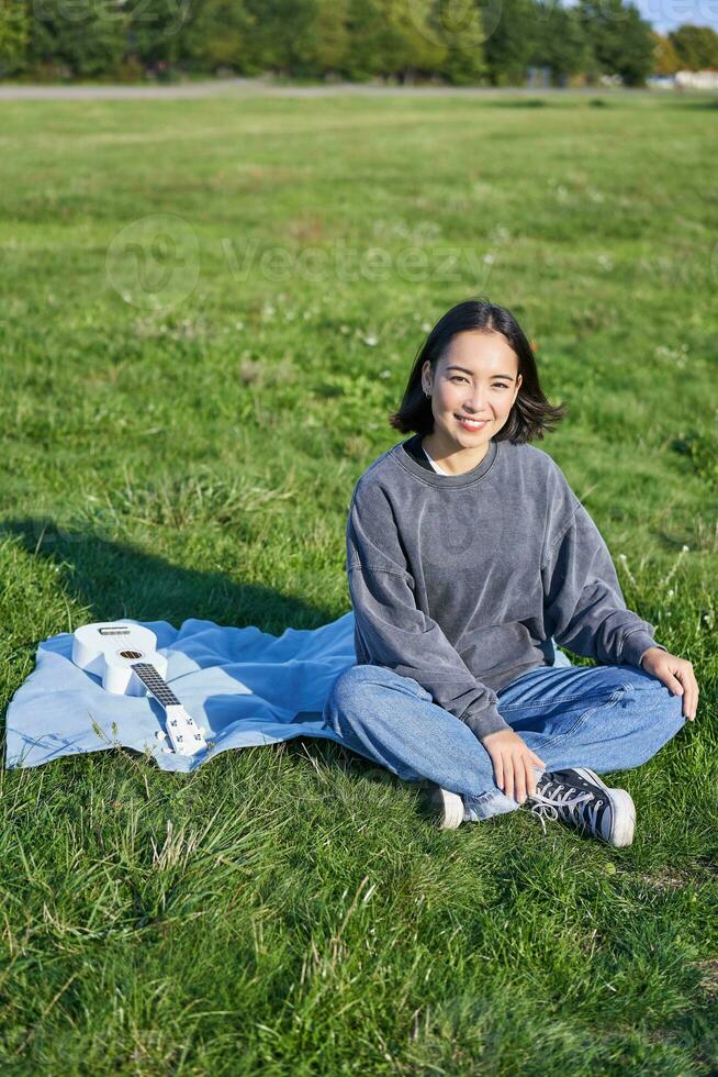 verticale coup de souriant asiatique fille séance sur couverture avec ukulélé guitare, relaxant sur ensoleillé journée en plein air, repos dans parc photo