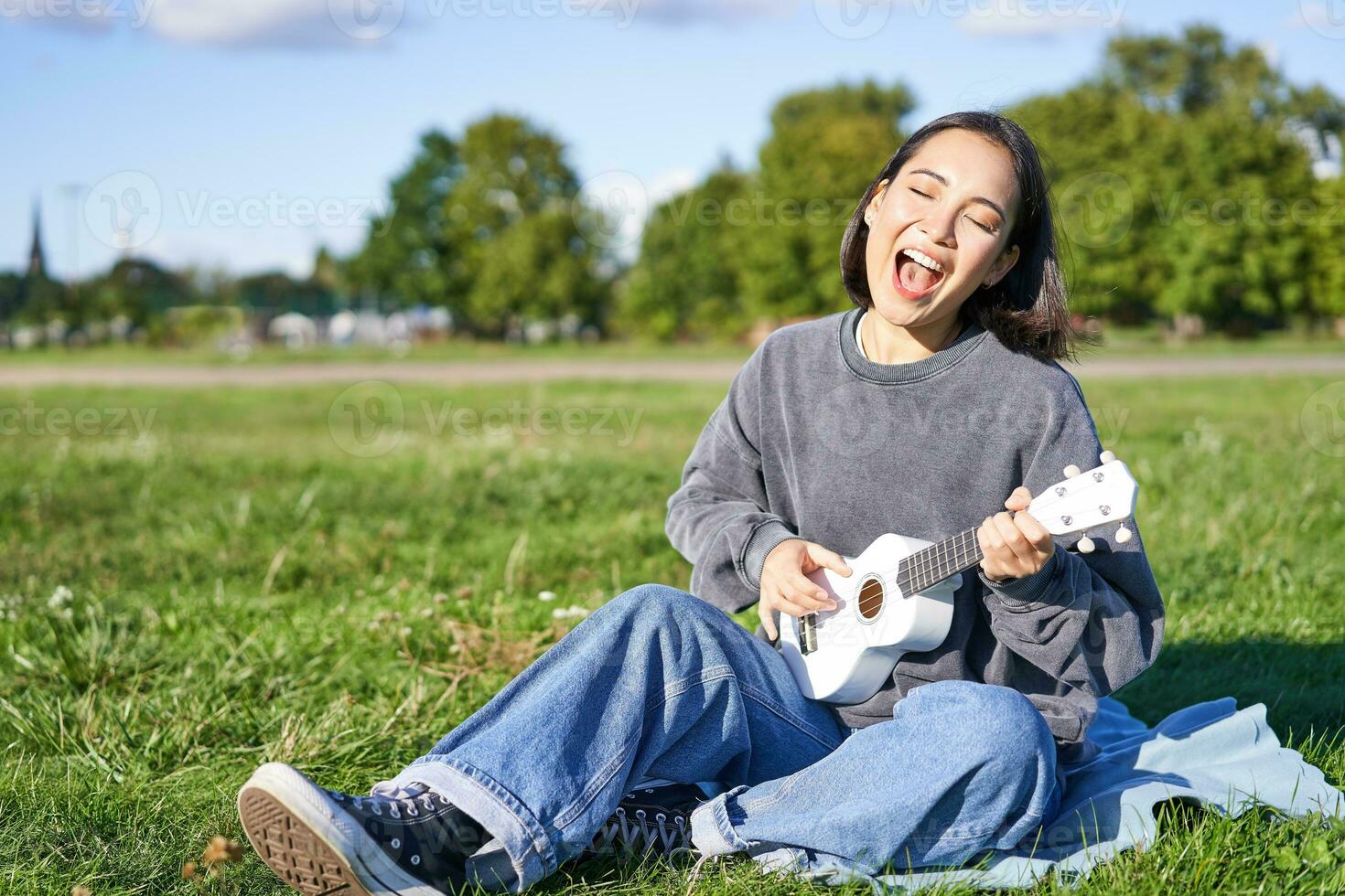 portrait de magnifique asiatique femme en chantant, en jouant ukulélé guitare dans parc, séance seul sur herbe sur ensoleillé journée photo