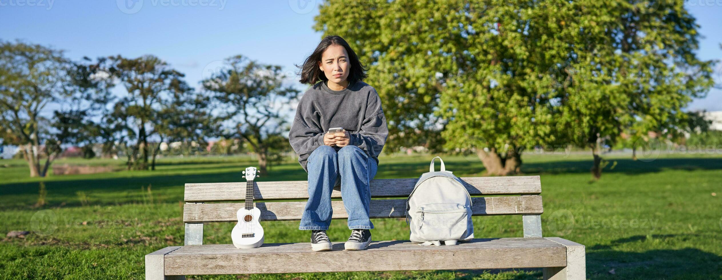 magnifique brunette fille sur banc dans parc, séance avec ukulélé et sac à dos, en portant téléphone intelligent, en utilisant mobile app photo