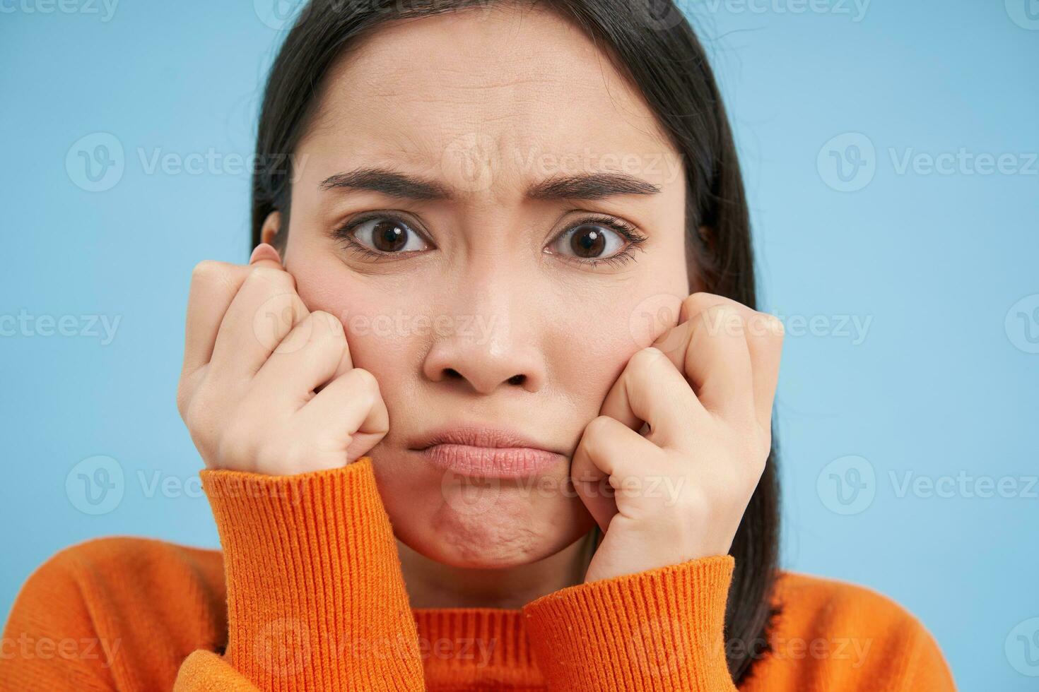 proche en haut de en colère bouder asiatique femme, faire la moue et renfrogné, en portant mains sur joues, se sent offensé, des stands plus de bleu Contexte photo