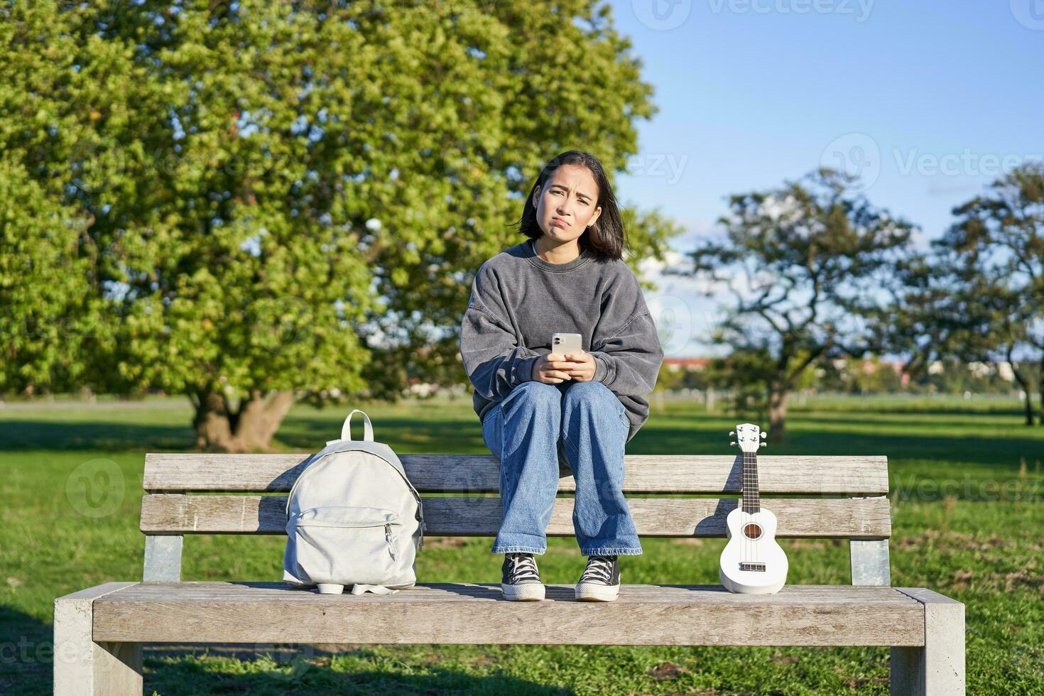 fille avec triste affronter, séance sur banc avec téléphone intelligent et ukulélé, à la recherche dérangé et déçu, étant seul dans parc photo