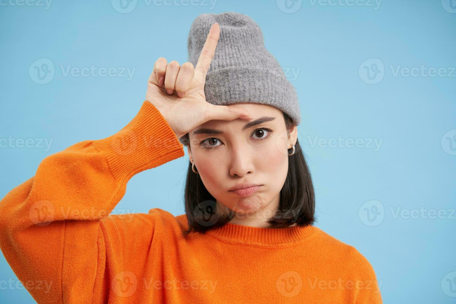 proche en haut portrait de asiatique fille étudiant dans bonnet, spectacles l lettre, perdant signe sur front, des stands plus de bleu Contexte photo