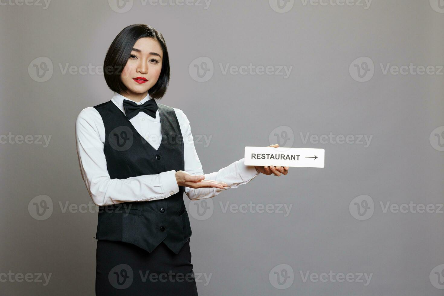 asiatique serveuse en portant restaurant direction signalisation et à la recherche à caméra tandis que posant dans studio. hospitalité un service femme employé dans uniforme montrant cafétéria aiguille acier signe portrait photo