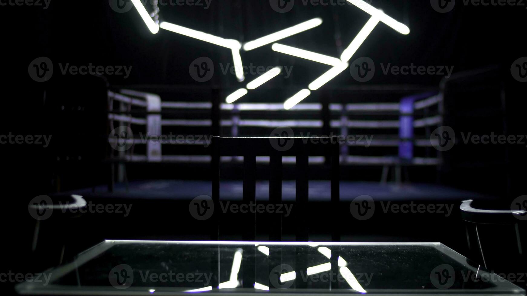 boxe bague et deux chaises avec table foncé Contexte. vue de une ordinaire boxe bague entouré par bleu Cordes éclairé par une projecteur. lumière spectacle autour le boxe anneau. photo