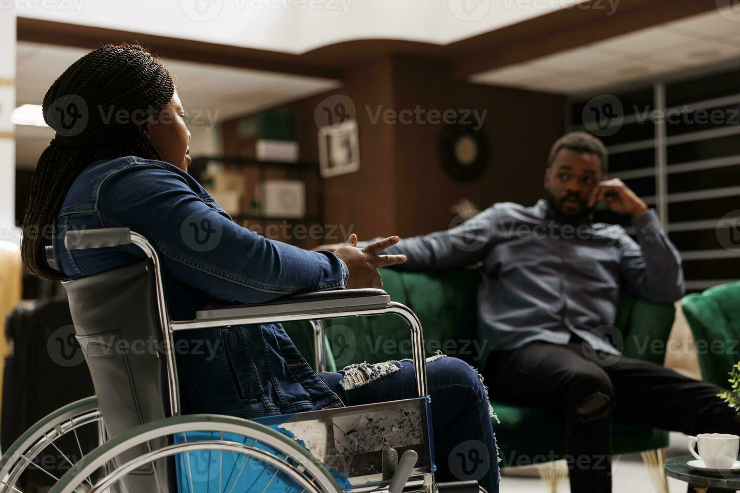 Jeune africain américain femme touristique avec invalidité séance dans Hôtel hall avec mari, en arrivant à fauteuil roulant accessible Hôtel. valide homme en voyageant avec désactivée petite amie photo