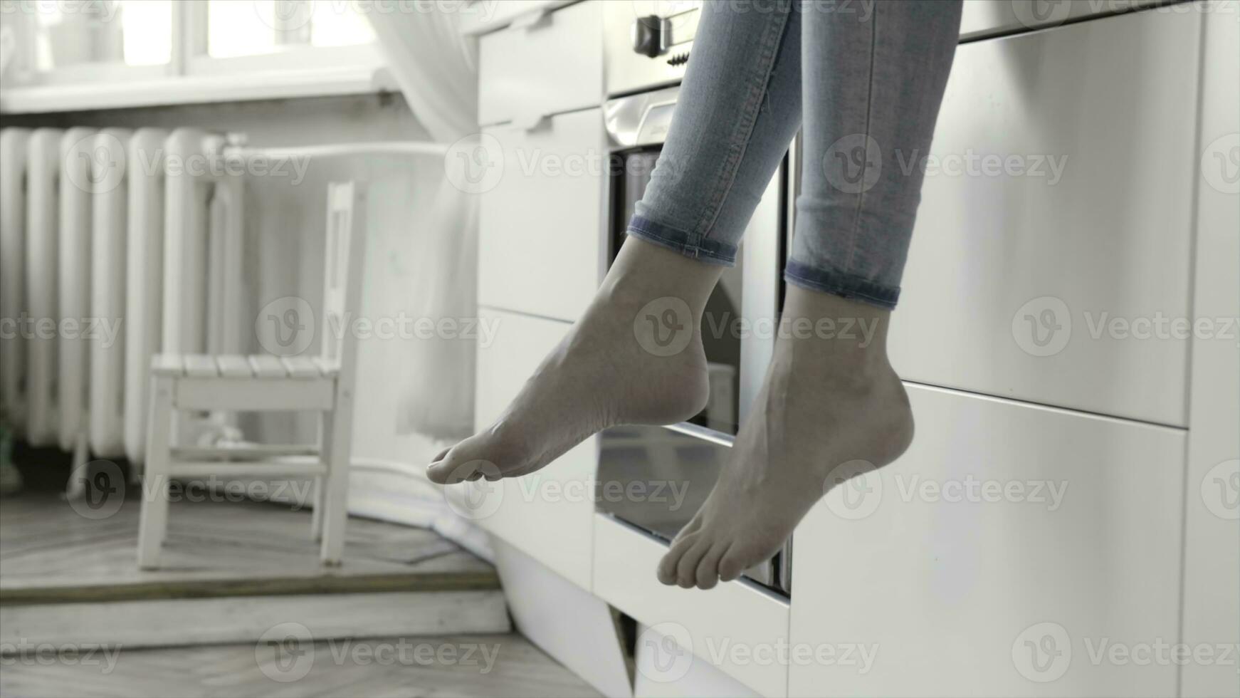 proche en haut pour femme jambes balançant avec blanc cuisine tiroirs et four sur le Contexte. action. femmes portant jeans séance pieds nus sur cuisine comptoir. photo