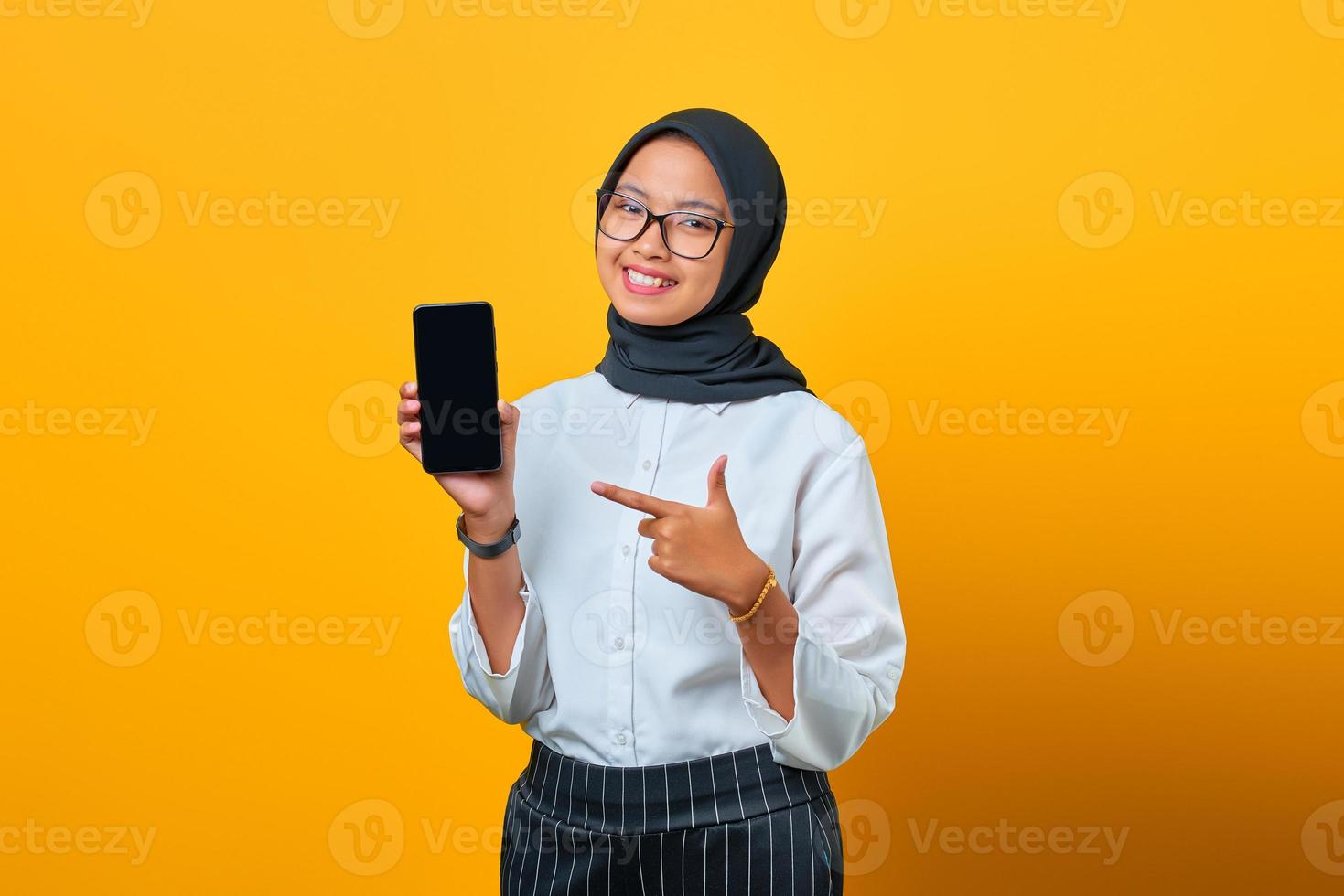 Happy young asian woman pointing at mobile phone écran blanc isolé sur fond jaune photo