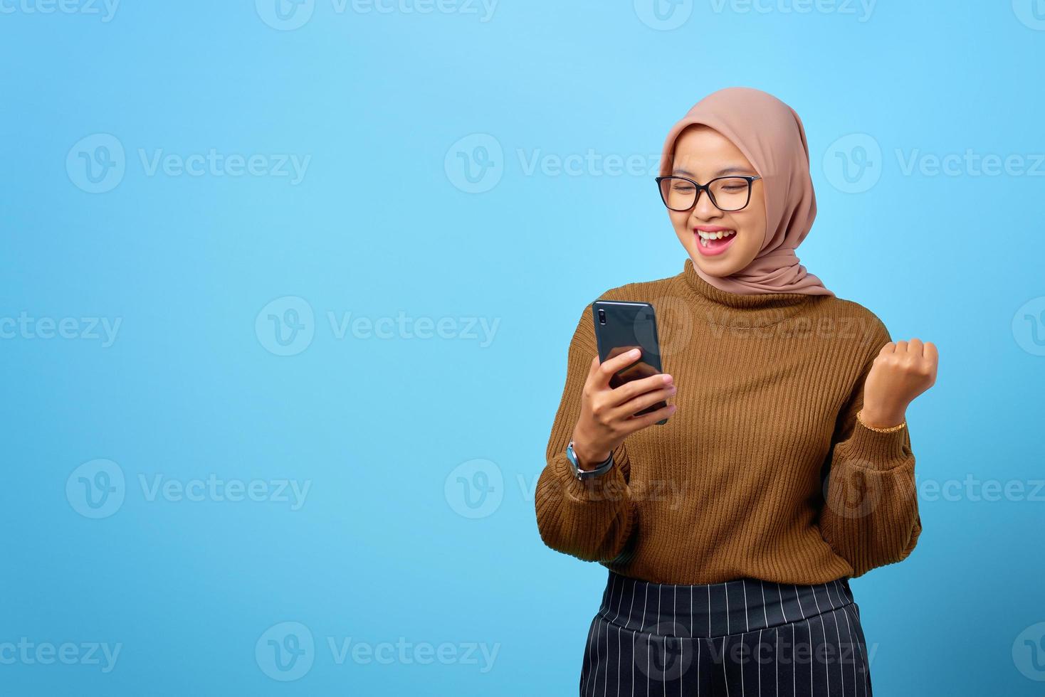 heureuse jeune femme asiatique utilisant un téléphone portable avec succès de geste de la main sur fond bleu photo