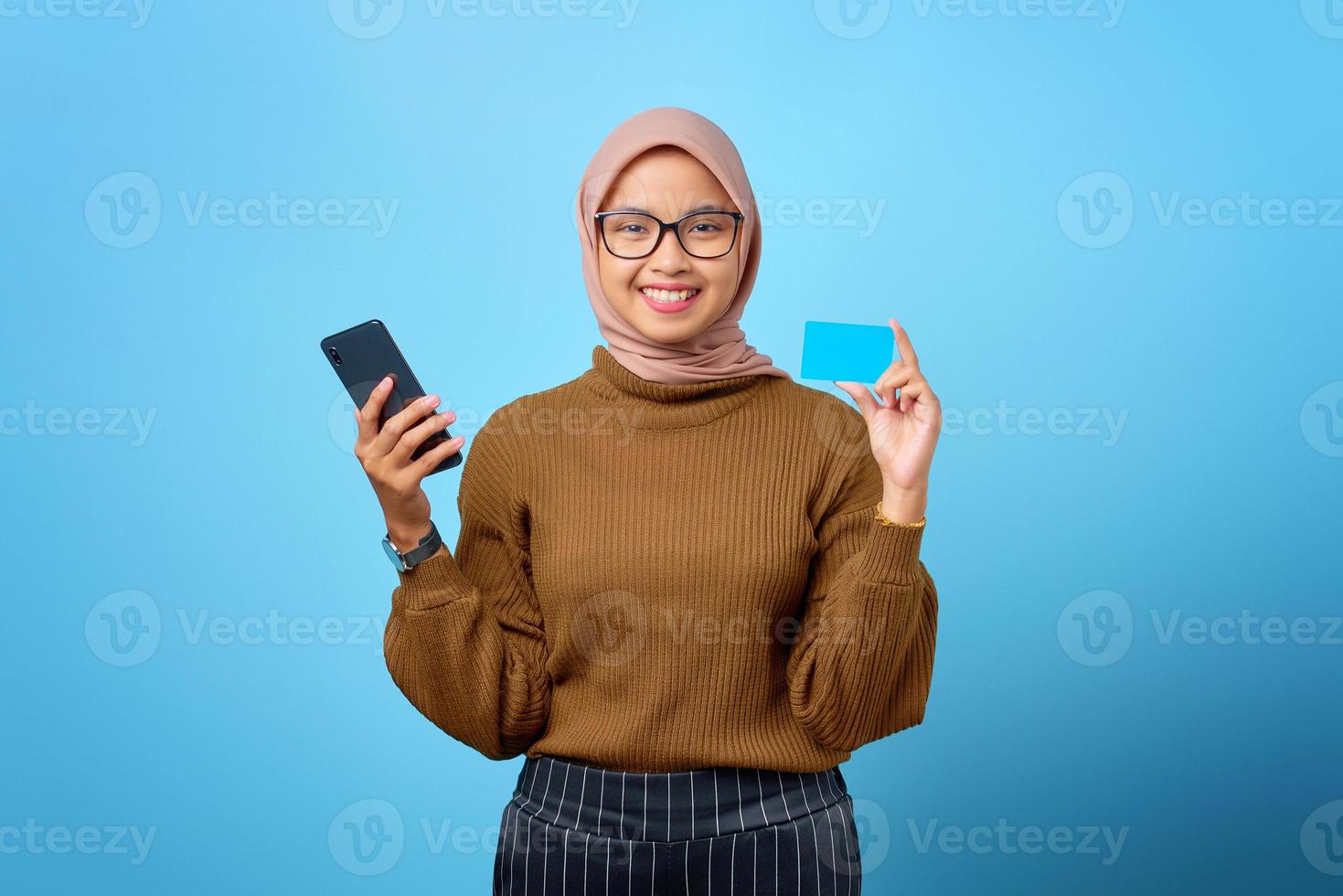 joyeuse jeune femme asiatique tenant un téléphone portable et une carte de crédit sur fond bleu photo