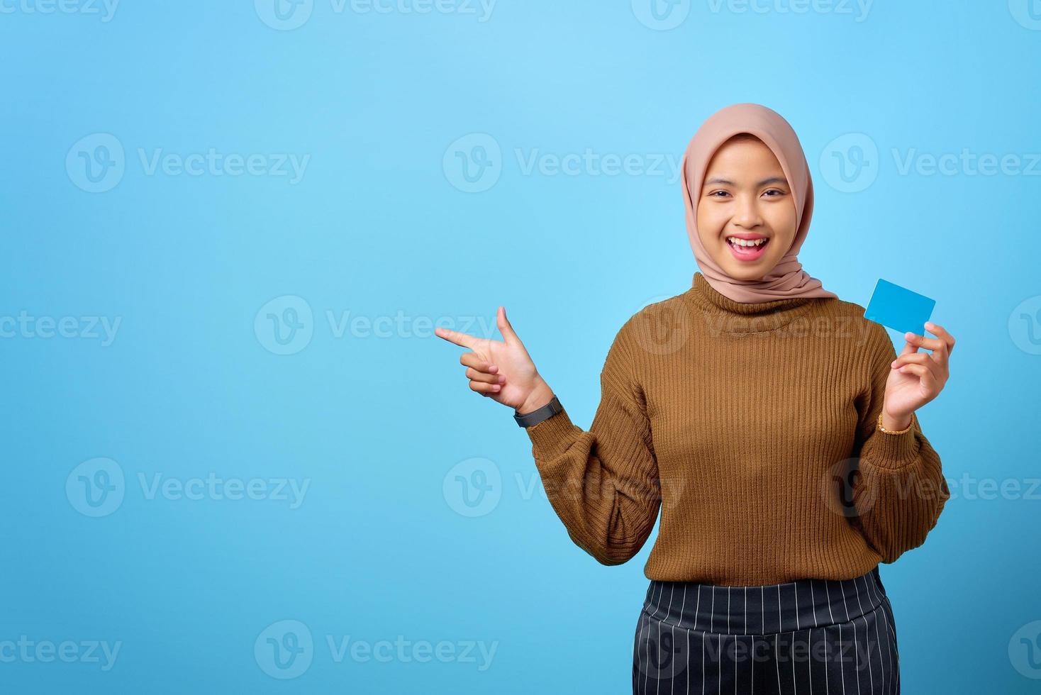 heureuse jeune femme asiatique tenant une carte de crédit et pointant du doigt pour copier l'espace sur fond bleu photo