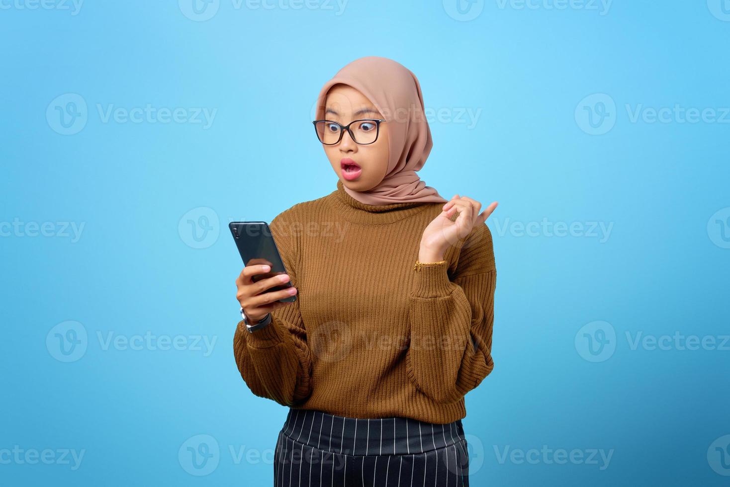 surpris jeune femme asiatique regardant l'écran du smartphone mobile avec la bouche ouverte sur fond bleu photo