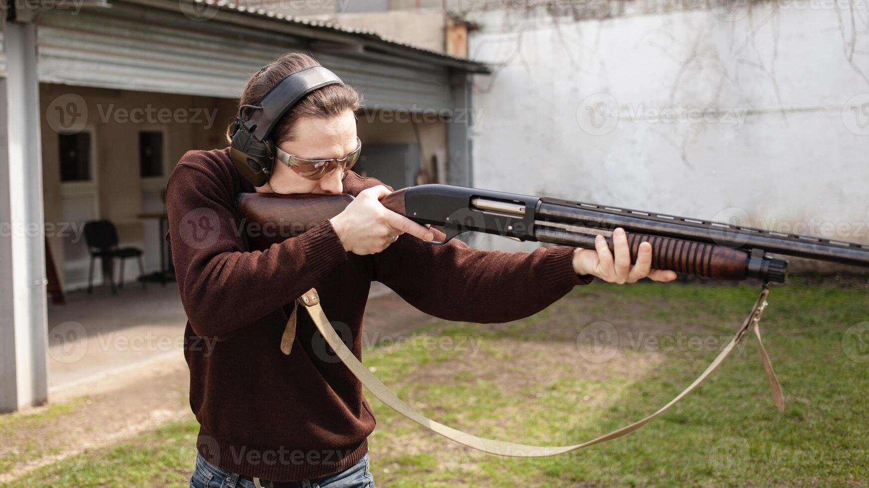 une homme avec une pompeux arme à feu sur une blanc Contexte. une homme dans protecteur des lunettes et écouteurs. pneu Extérieur. gris hangar. pistolet photo