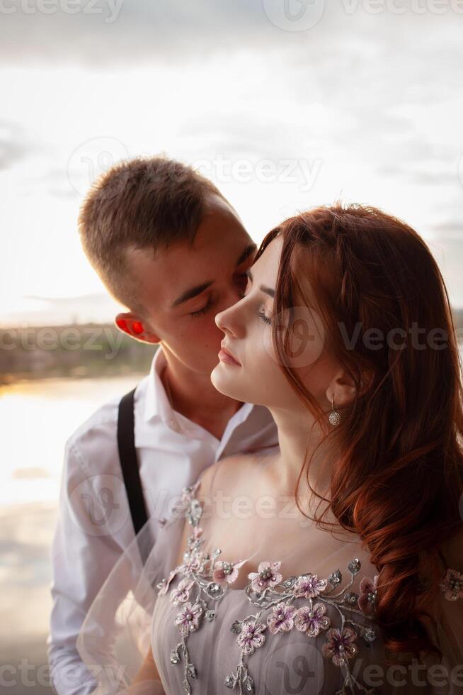 une magnifique mariage couple sur le berge de rivière à le coucher du soleil. une femme dans une gris robe avec fleurs, une homme dans une costume et bretelles étreinte chaque autre, l'amour. les amoureux sur une marcher photo