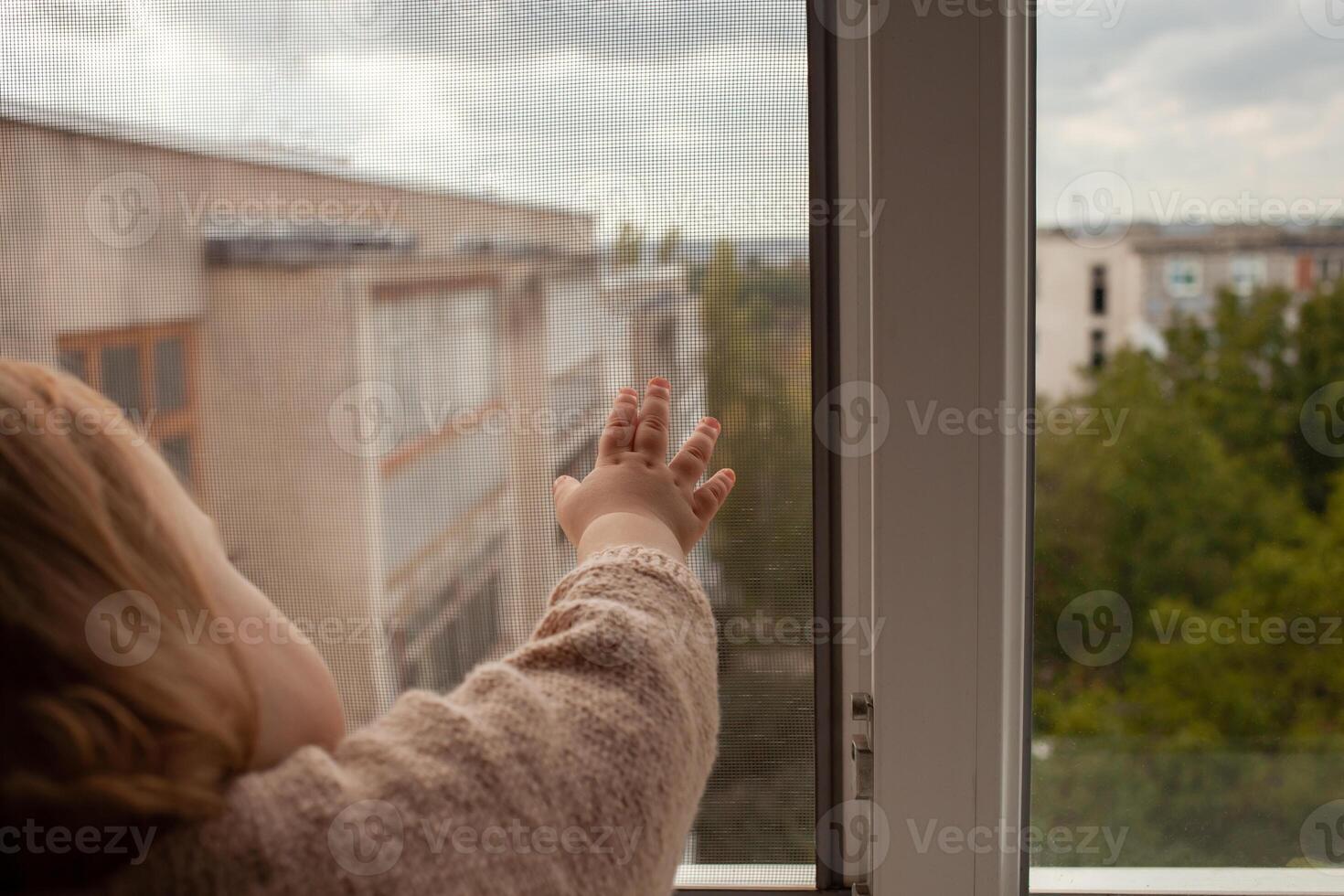 le enfant grimpe à le fenêtre, le fille sur le fenêtre seuil repose sur le filet, le danger de chute. le enfant est seul à maison, pouvez tomber en dehors de le la fenêtre. photo