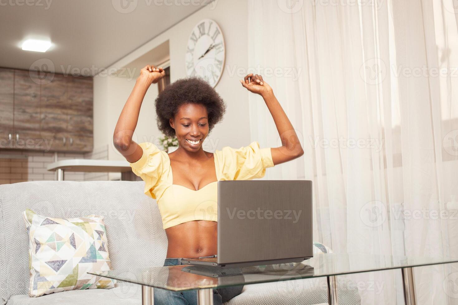 une africain américain femme pigiste, travail de maison. femme dans de face de portable est content, souriant. gagnant loterie, embauché, les stocks avoir ressuscité, crypto-monnaie a grandi, bien nouvelles. surpris photo