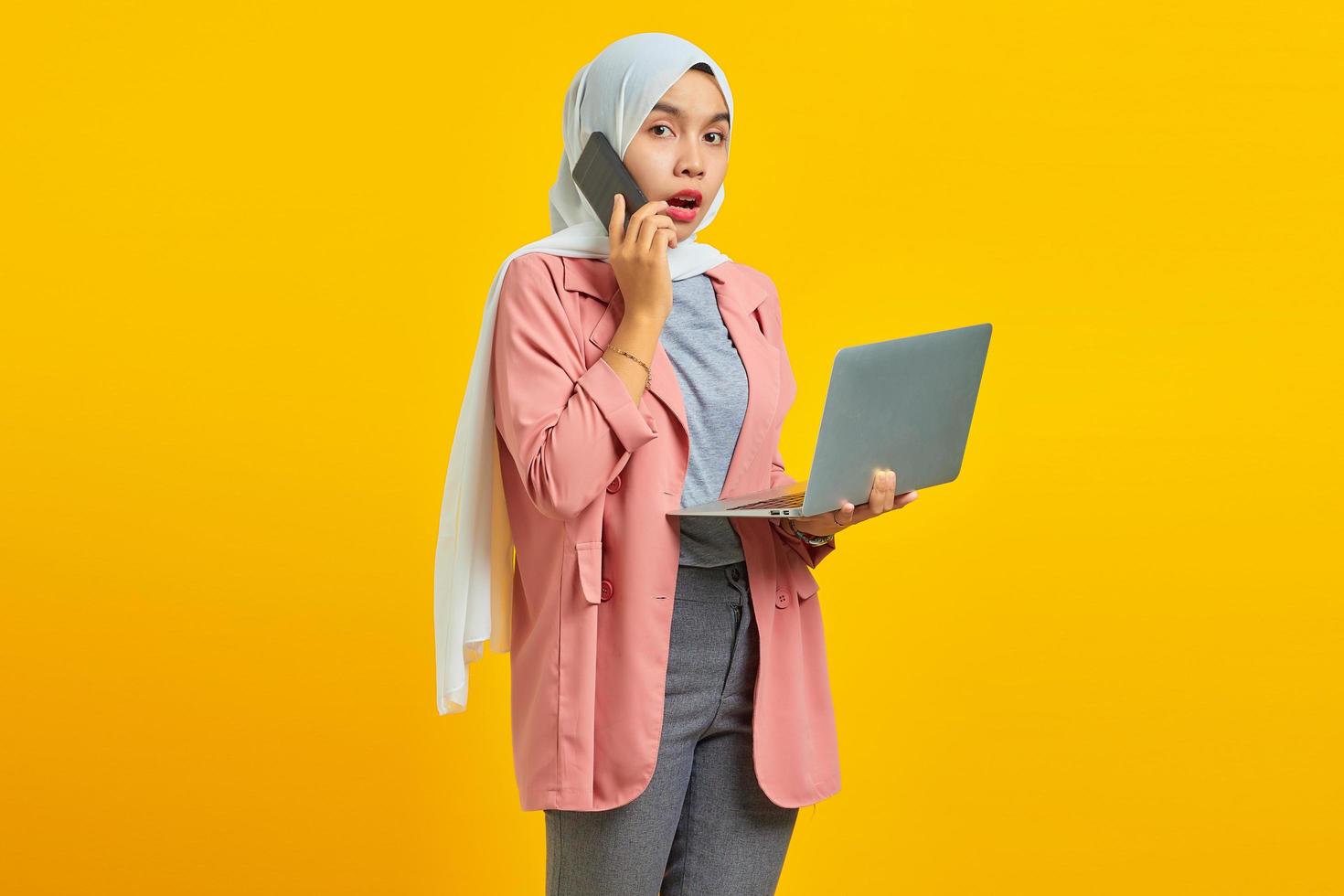 portrait d'une femme asiatique surprise et choquée parlant sur un smartphone tout en tenant un ordinateur portable argenté isolé sur fond jaune photo